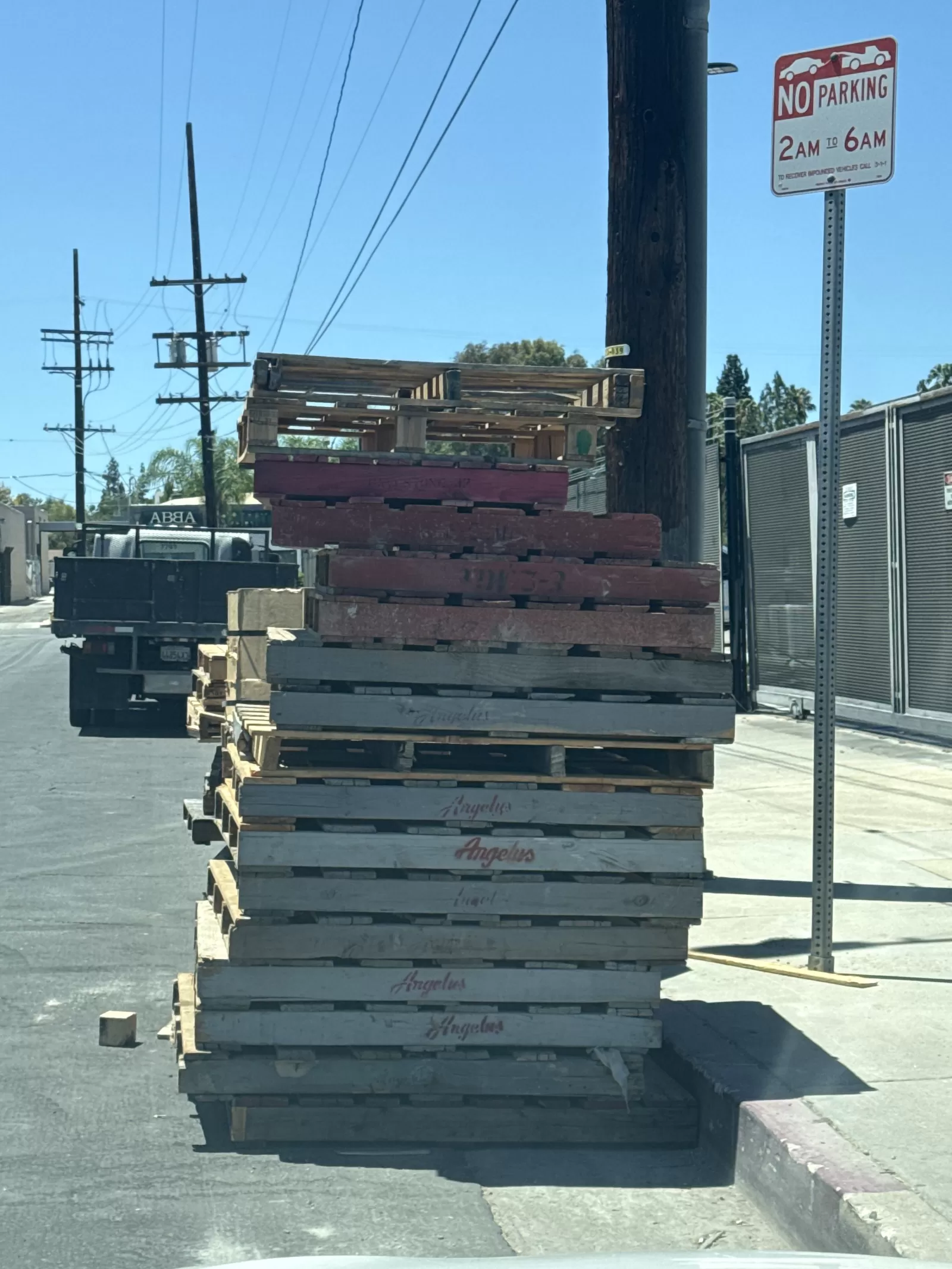 A large stack of wooden pallets occupies the curbside roadway and extends beside the sidewalk. A small loose block is also visible on the pavement nearby.